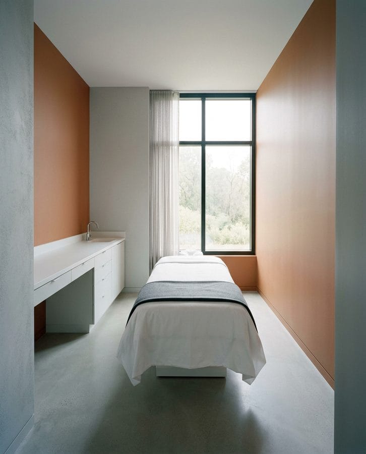 A tall view into an empty treatment room with a flat bed in fresh linen and a window at the far end.