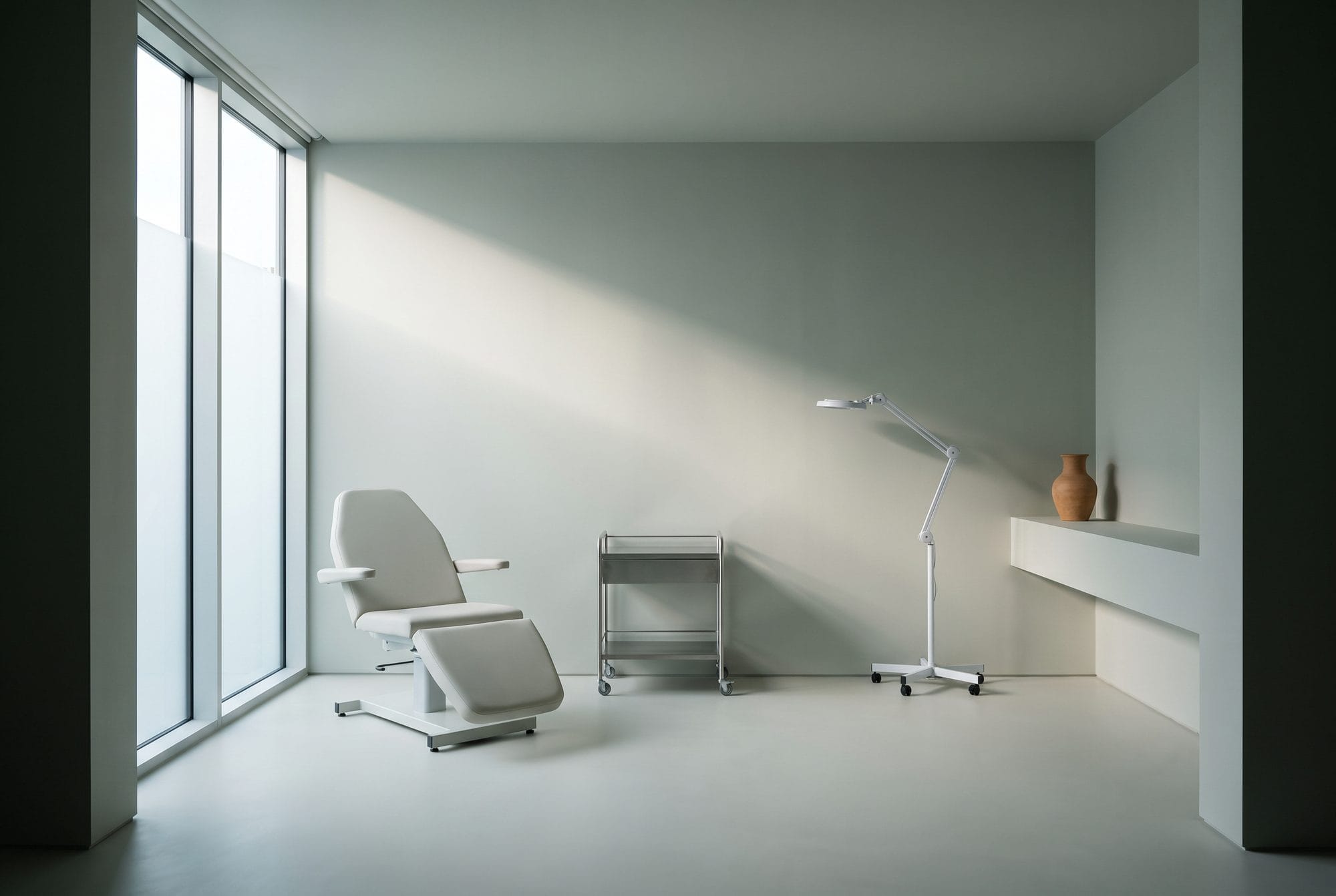 An empty treatment room in morning light, a plain adjustable chair set at an angle beside a full-height window.