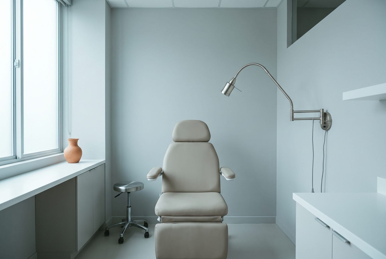 An empty treatment room with a plain adjustable chair, a rolling stool and a wall-mounted articulated light.